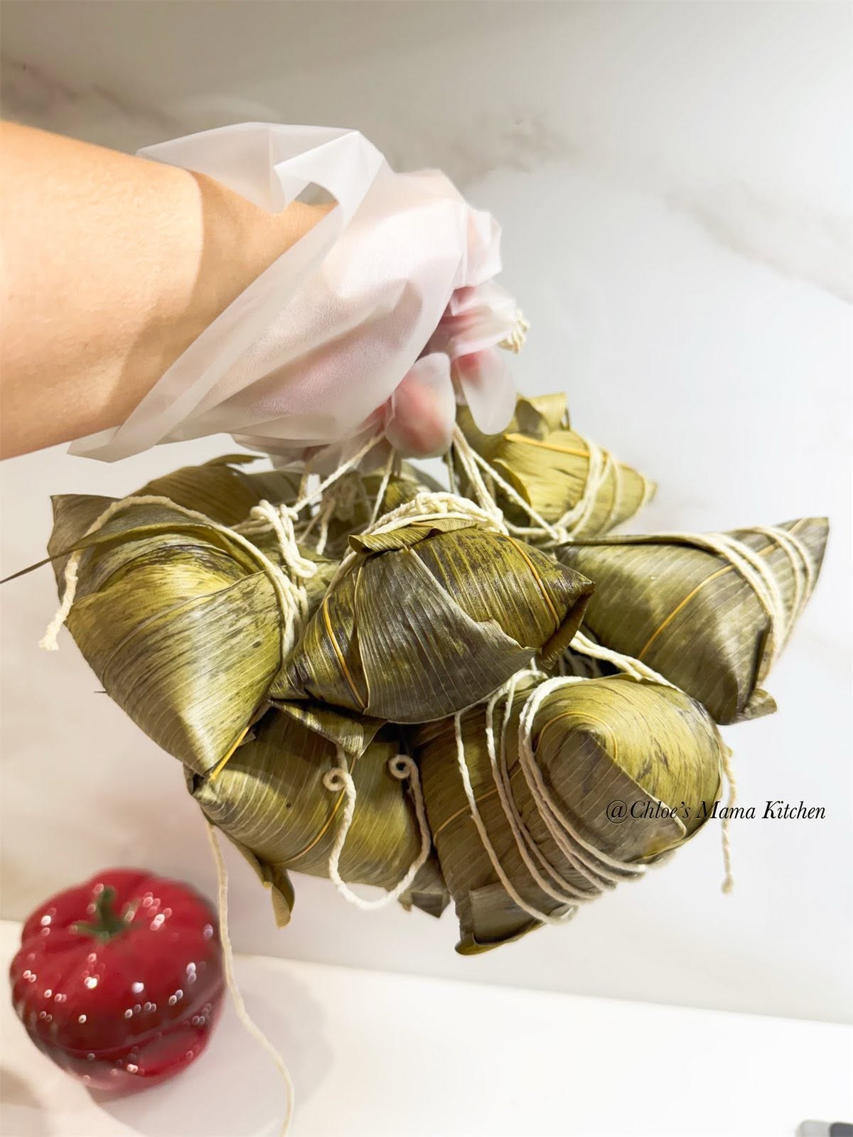 A gloved hand holding Chloe's Mama Kitchen leaf-wrapped Bak Zang rice dumplings tied with string.