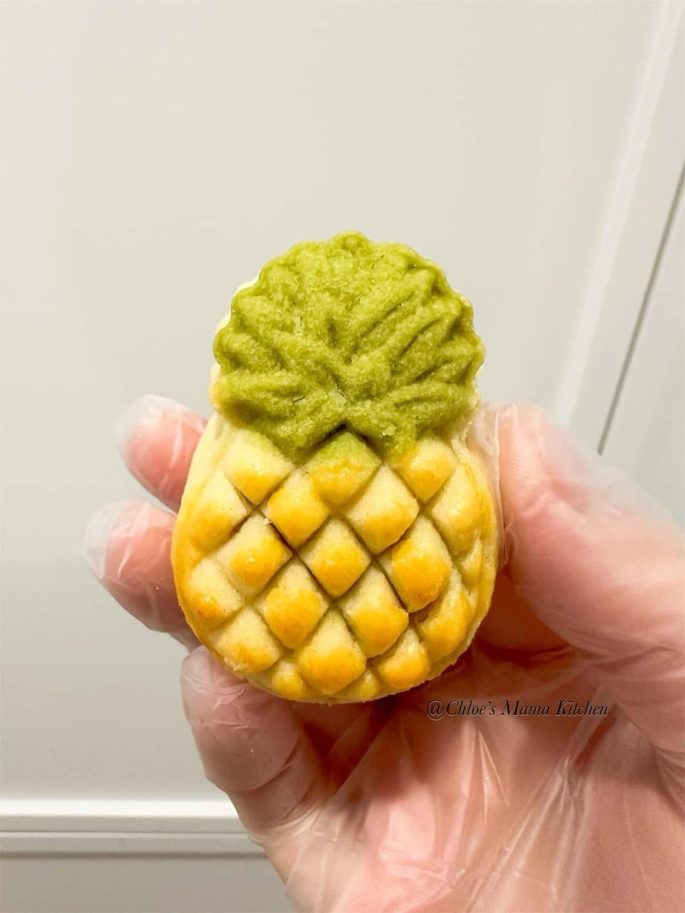 A handmade pineapple tart with golden pastry and green matcha leaves, held in a gloved hand
