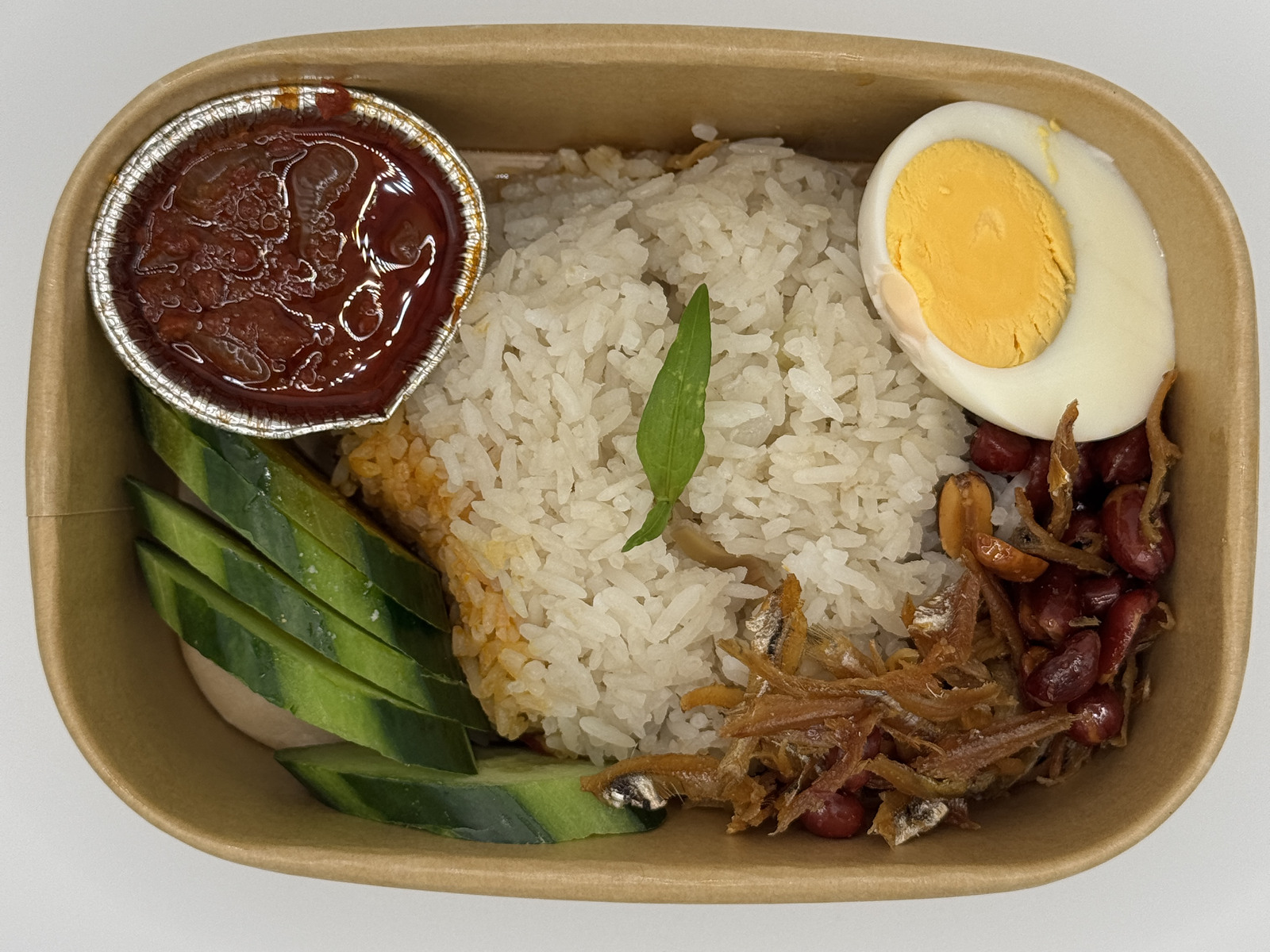Malaysian-style Nasi Lemak with coconut rice, sambal, egg, cucumber, peanuts and anchovies in a takeaway box.