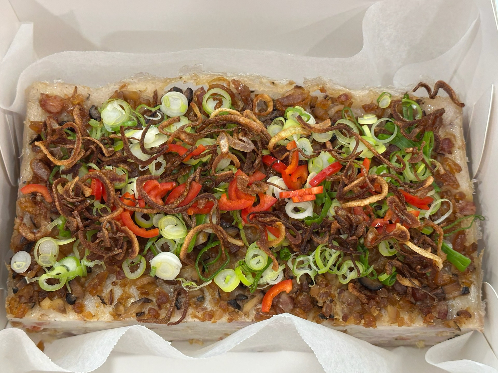 Taro Cake in a paper box, topped with Chinese sausage, spring onion, chilli and fried shallots. Photograph does not establish the selling portion.